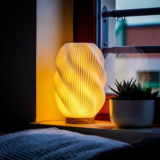 Helix 3D printed table lamp with spiral design and oak wood base glowing warmly on a window sill.