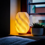 Helix 3D printed table lamp with spiral design and oak wood base glowing warmly on a window sill.