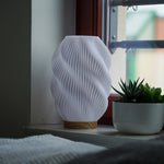 Helix 3D printed table lamp with spiral design and oak wood base glowing warmly on a window sill.