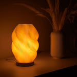 Helix 3D printed table lamp with spiral design and oak wood base glowing warmly on a window sill.